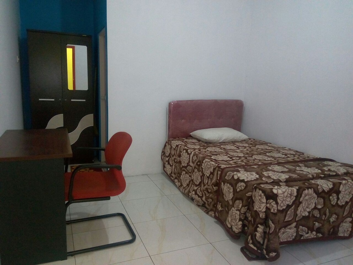 A comfortable room features a single bed with a patterned blanket, complemented by a desk and a red chair. A cabinet is positioned against the wall, and a door leads to another space, providing privacy and access.