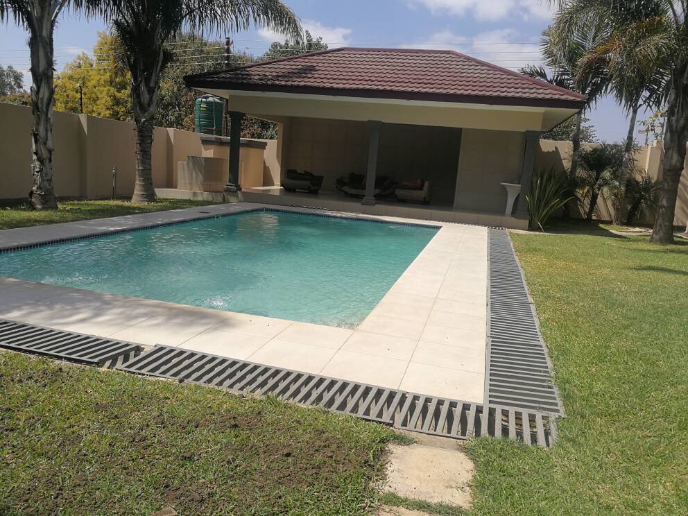 A clear swimming pool is framed by neatly arranged tiles and surrounded by well-maintained grassy areas. In the background, a covered seating area is visible, offering shaded space for relaxation under palm trees.