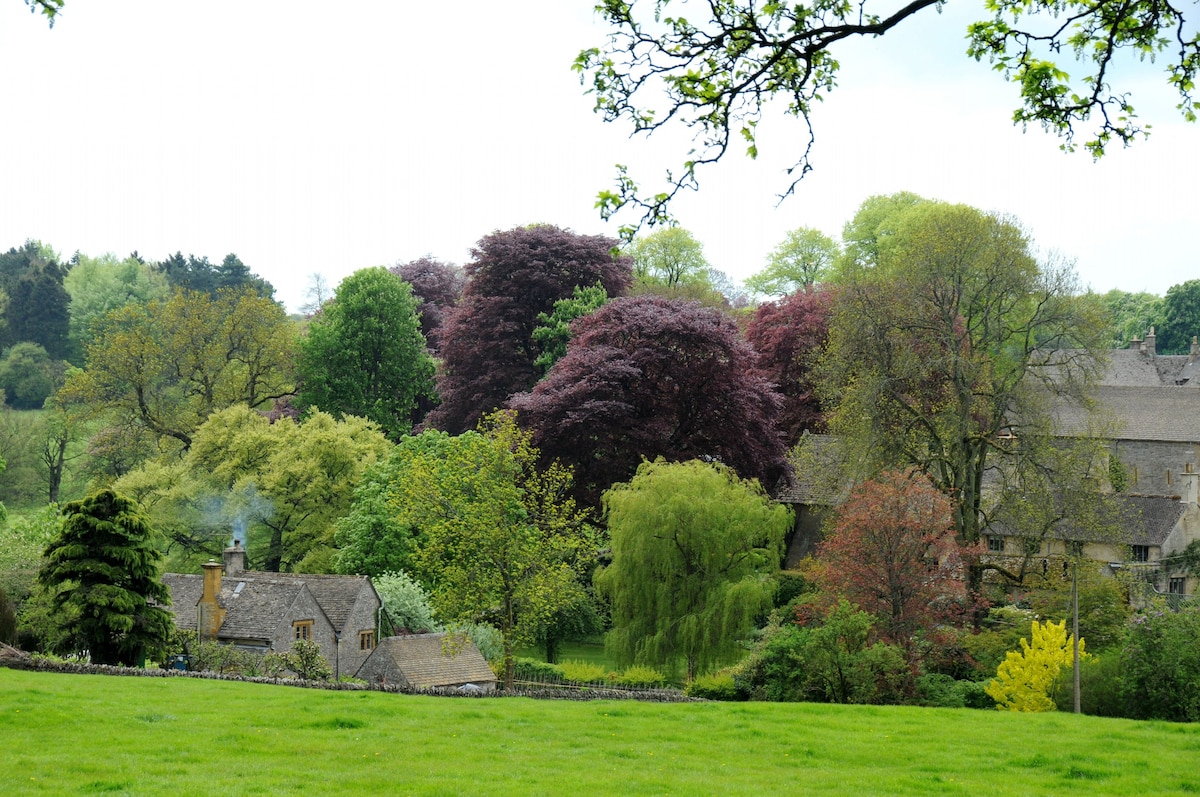 A serene countryside scene showcases a patchwork of trees in varying shades of green and purple, providing a vibrant backdrop. A quaint stone cottage gently rises amidst the landscape, with smoke softly billowing from its chimney. Rolling hills invite exploration and offer a sense of tranquility.