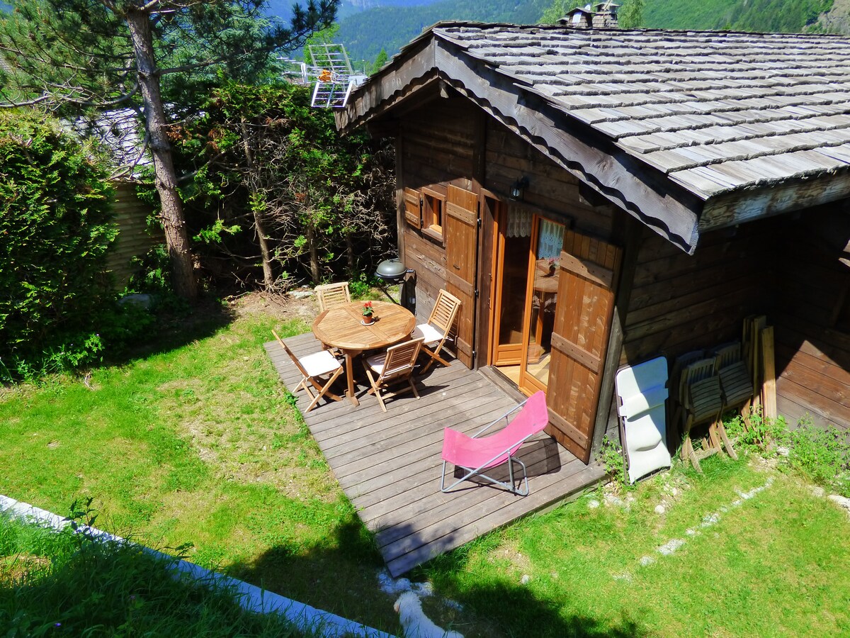 A wooden chalet is set in a green outdoors area, featuring a small terrace with a round table and four chairs. A pink lounge chair is positioned nearby. The chalet’s double doors open to the exterior, framed by trees and mountainous scenery in the background.