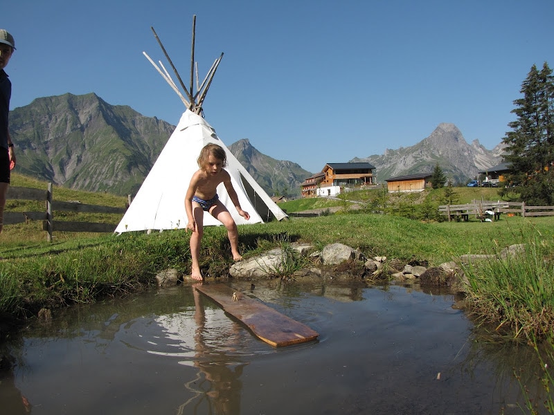 A child is seen balancing on a wooden plank above a shallow pond in a serene outdoor setting. A teepee stands in the background, surrounded by lush greenery. Majestic mountains rise in the distance under clear blue skies, enhancing the natural landscape.