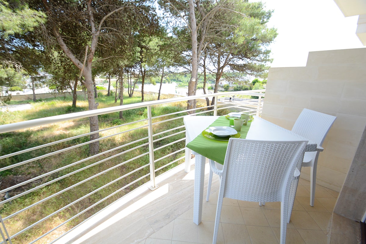 A balcony equipped with a small dining table and chairs is set against a backdrop of trees and greenery. The table is adorned with a green tablecloth, providing a space for outdoor dining while enjoying views of the surrounding landscape.