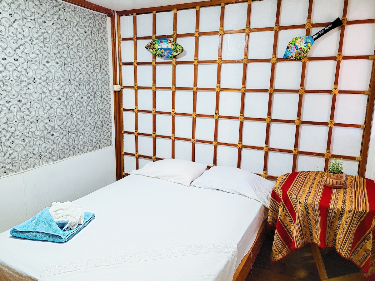 A private room features a spacious bed with a white covering. Textured wooden panels create a unique backdrop, accompanied by decorative art pieces. A small table adorned with a colorful cloth and a plant complements the space, creating a serene and comfortable environment.