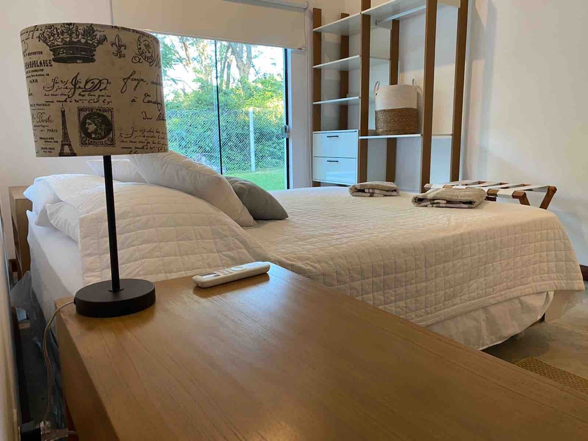 A well-lit bedroom features a spacious bed with a neutral quilted cover. A lamp with a patterned shade sits on a wooden nightstand, while decorative towels are neatly arranged. Natural light enters through a large window, overlooking a green outdoor space.