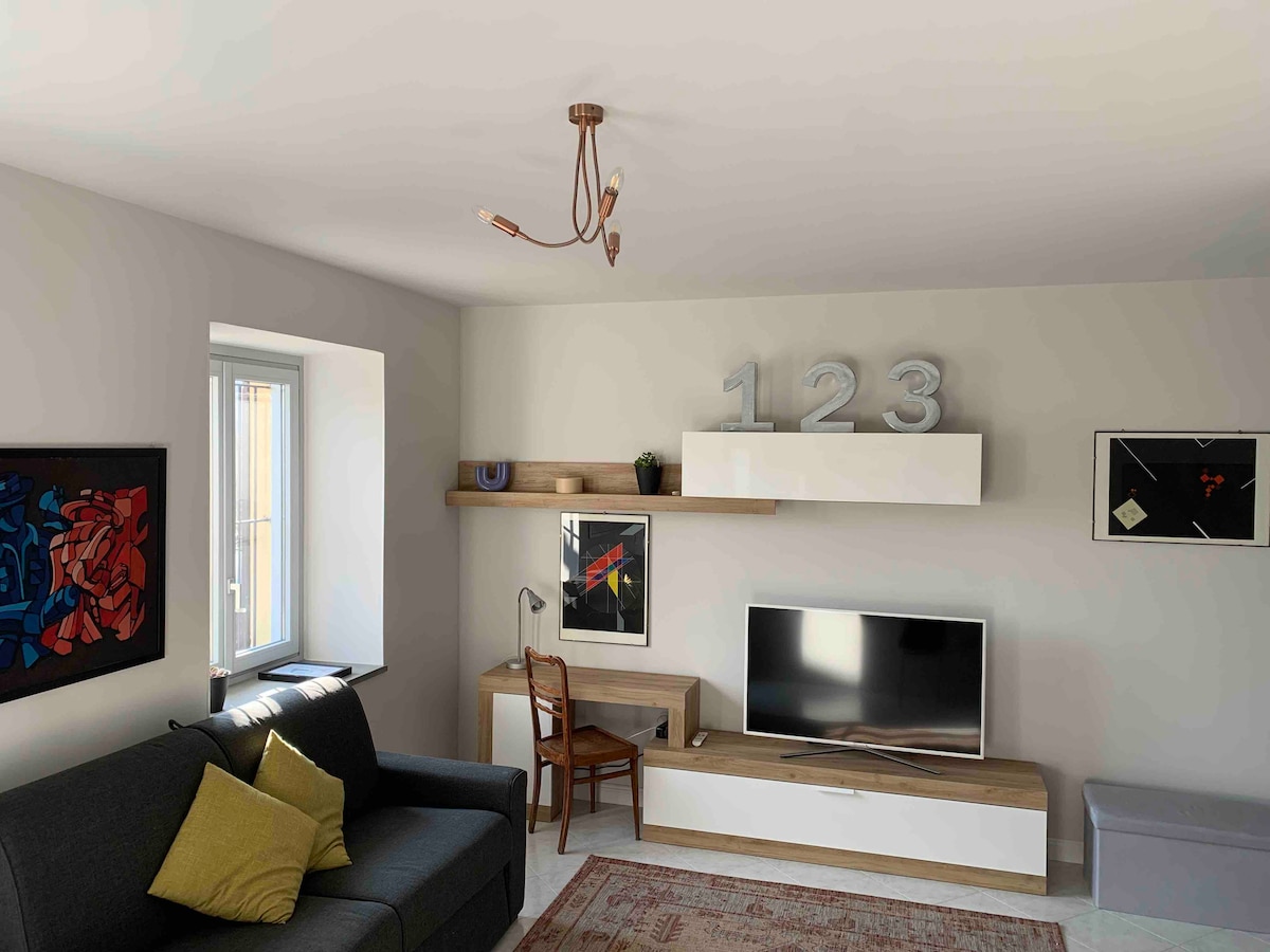 A modern living area features a comfortable dark sofa with a green pillow. A wooden shelf holds decorative items, while a TV unit provides storage beneath a wall-mounted television. Large windows allow natural light to brighten the space, complemented by colorful artwork on the walls.