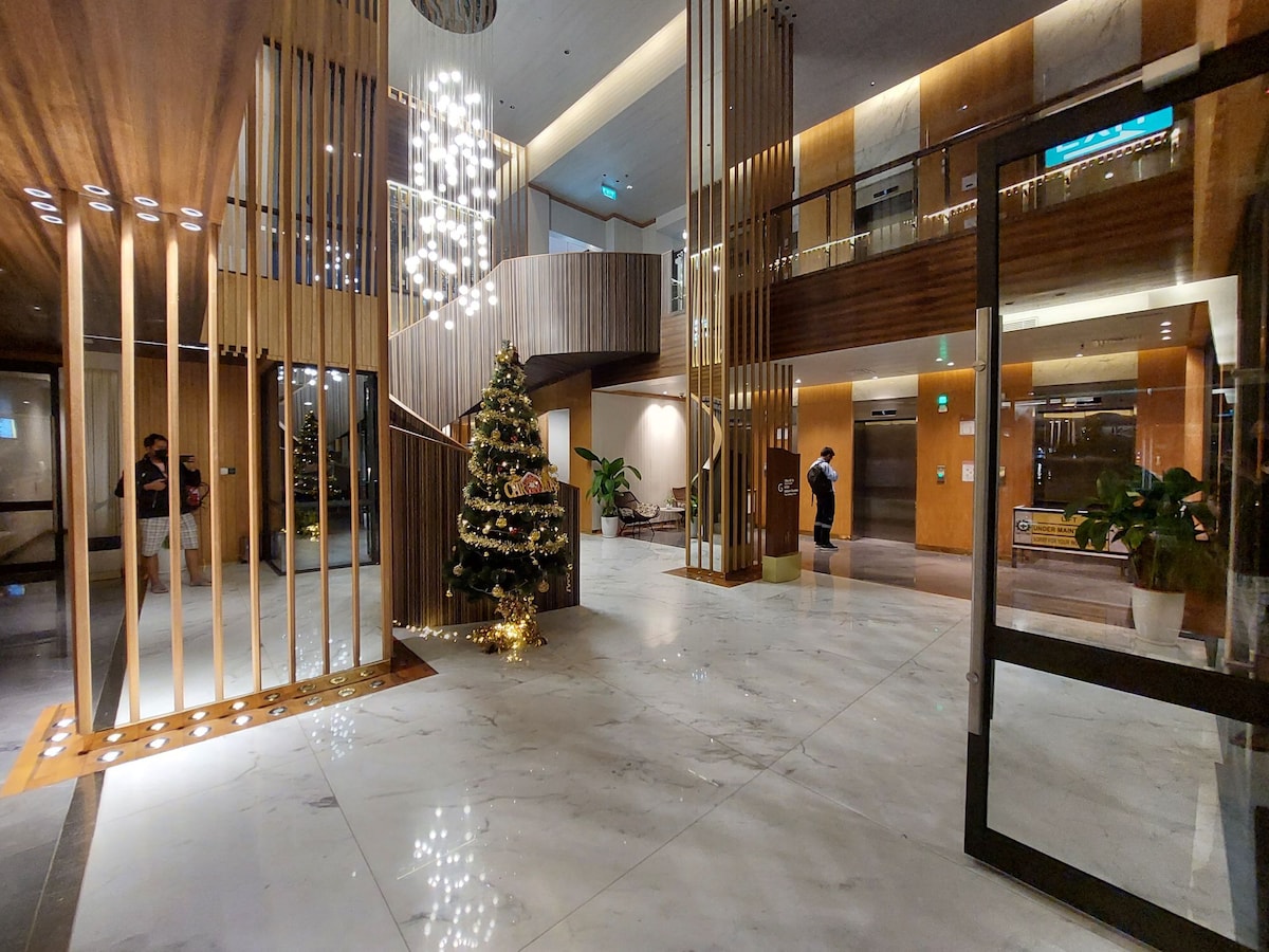 An elegant lobby is shown with wooden accents and soft lighting. A decorated Christmas tree stands prominently in the corner, surrounded by warm light. The space is complemented by a series of vertical wooden dividers and a spacious staircase in the background.