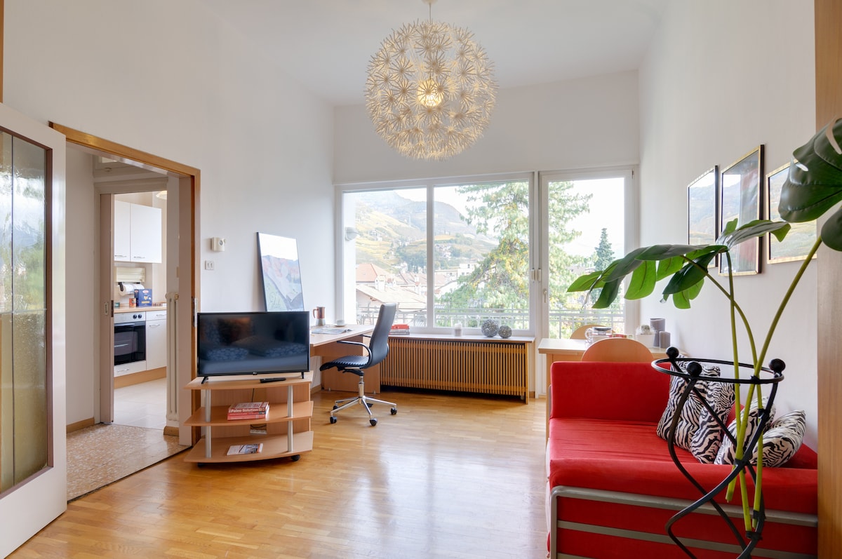A light-filled living area showcases a cozy seating space with a red couch and a decorative plant. A modern workspace is positioned by a large window, offering scenic views. The room features a contemporary light fixture and subtle wooden flooring, contributing to a bright and open ambiance.