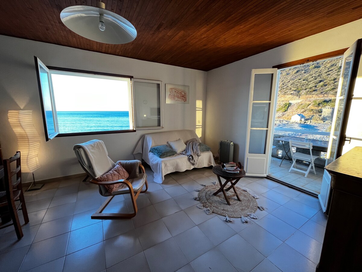 A light-filled living space features large windows offering views of the sea. A cozy white sofa sits beside a round coffee table on a textured rug. Wooden chairs and a floor lamp create a welcoming ambiance. The interior design reflects a serene coastal style.