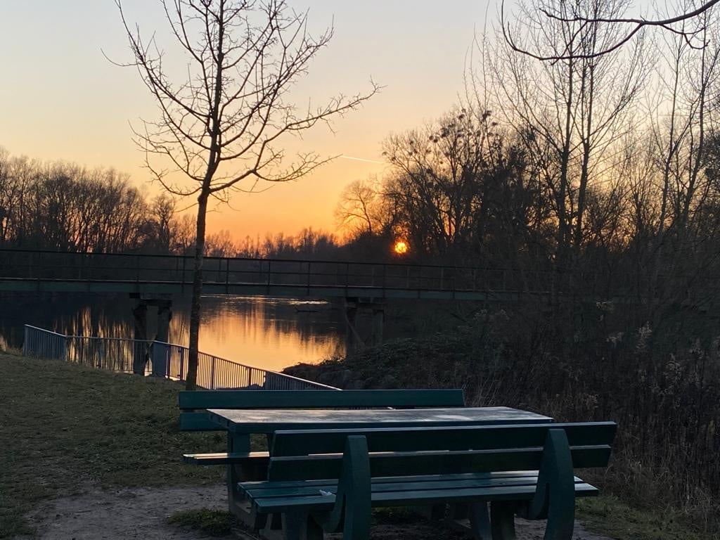 A quiet riverside scene is depicted at sunset, showcasing a simple green picnic table set by the water's edge. Silhouettes of trees frame the tranquil landscape, while the soft glow of the setting sun reflects off the water's surface, creating a serene atmosphere.