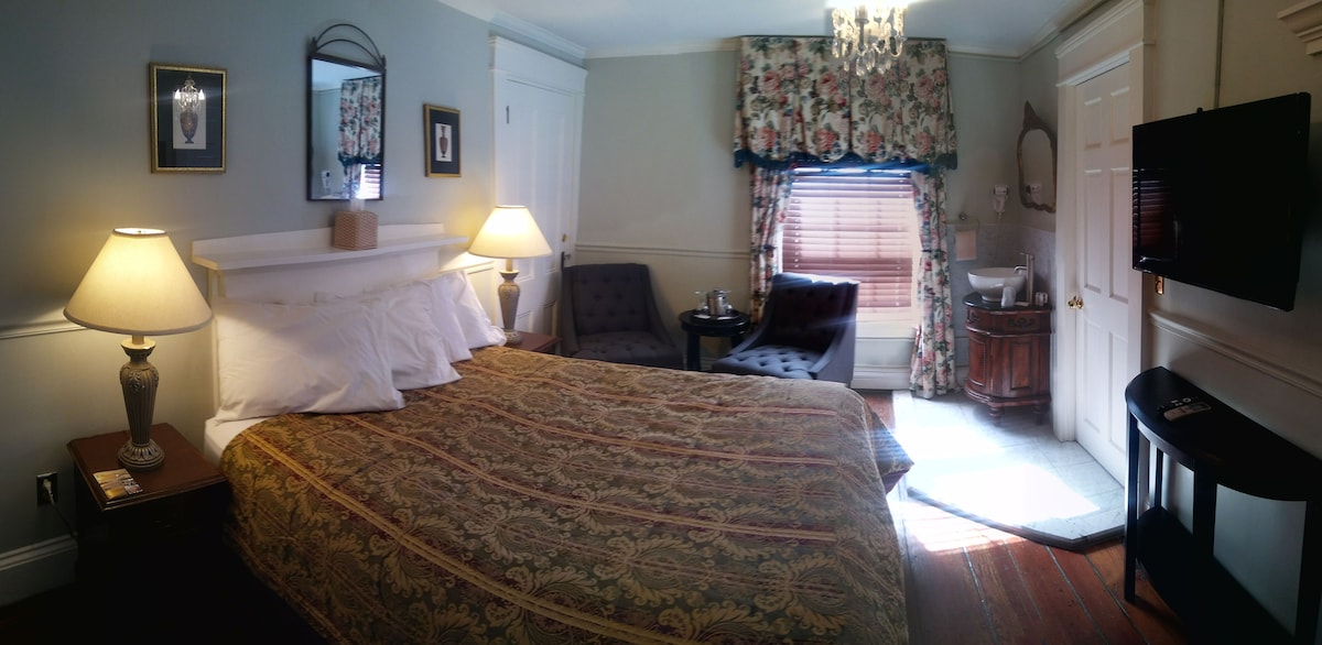 A well-appointed bedroom features a spacious bed with decorative bedding. Soft lighting from bedside lamps and a chandelier complements the floral drapes at the window. A seating area with two chairs invites relaxation, with a small table positioned nearby.