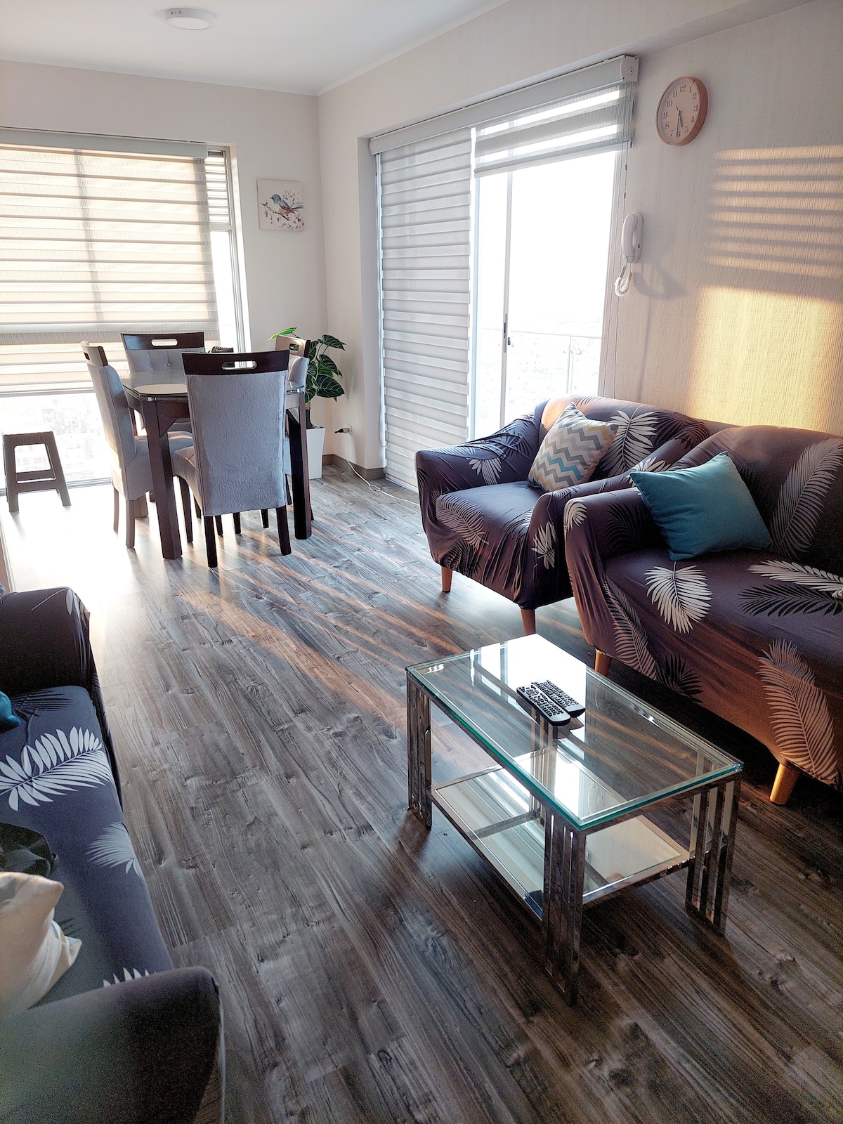 A modern living space features a glass coffee table at the center, surrounded by patterned sofas. Dining chairs and a table are visible in the background, while natural light streams through the large windows, highlighting the hardwood floor and decorative plant.