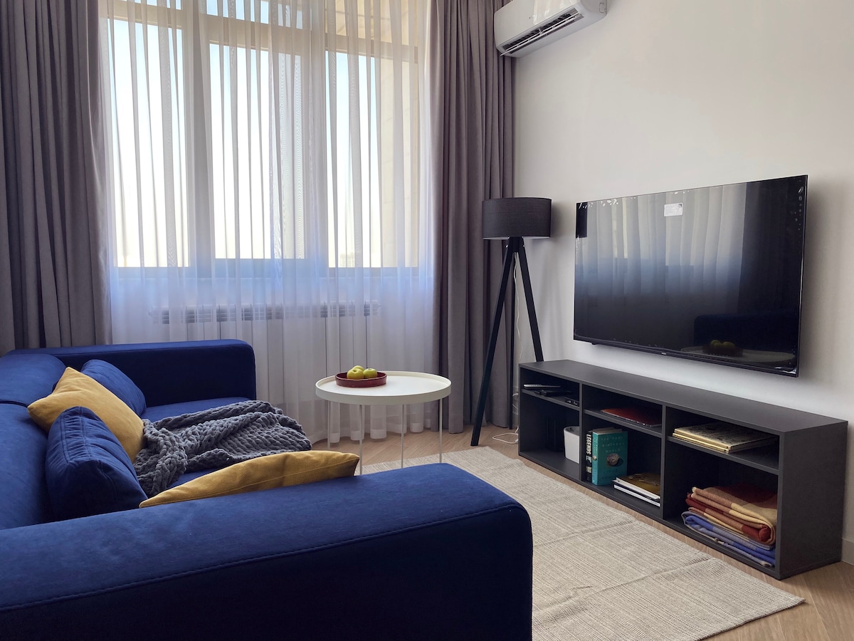 A modern lounge area features a deep blue sofa adorned with yellow cushions and a cozy blanket. A circular white coffee table is placed in front, while a large television is mounted on the wall. Natural light filters through sheer curtains, creating a bright atmosphere.