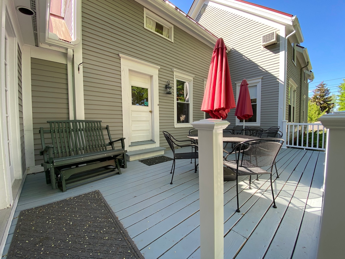 An outdoor deck features a green wooden bench and a black dining set. Two red umbrellas provide shade, while a light blue sky and greenery surround the area. The entrance door is visible, framed by light gray siding.
