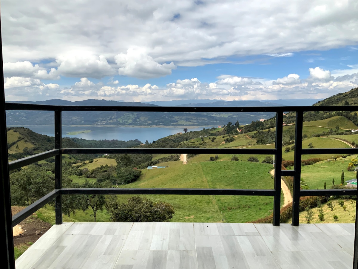A spacious balcony offers unobstructed views of the vast landscape, featuring rolling green hills and the shimmering surface of the Embalse de Tomine in the distance. A cloudy sky adds depth to the serene atmosphere.