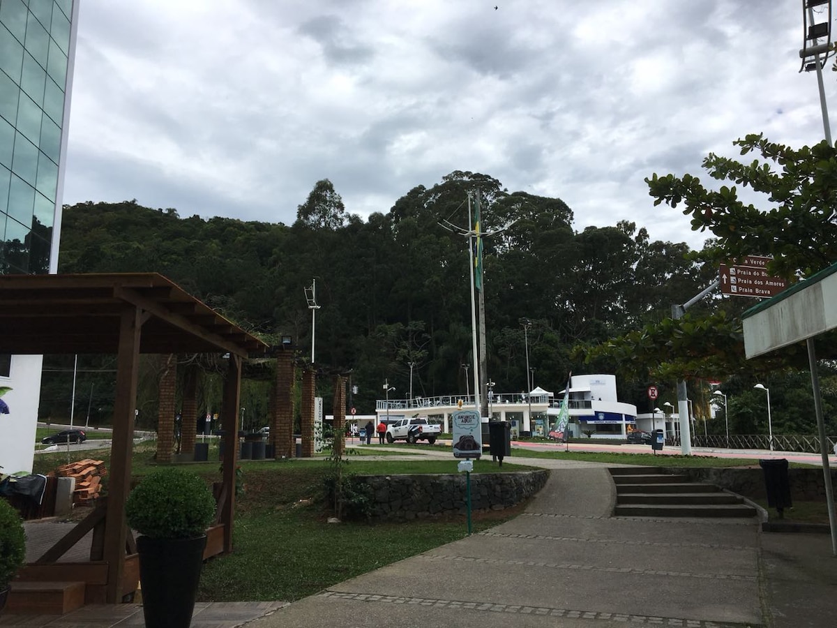 A landscaped outdoor area is visible, featuring a pathway leading towards a parking lot. Green trees and shrubbery line the pathway, with some wooden structures and benches visible. Overcast skies provide a subdued light, creating a calm and serene environment.