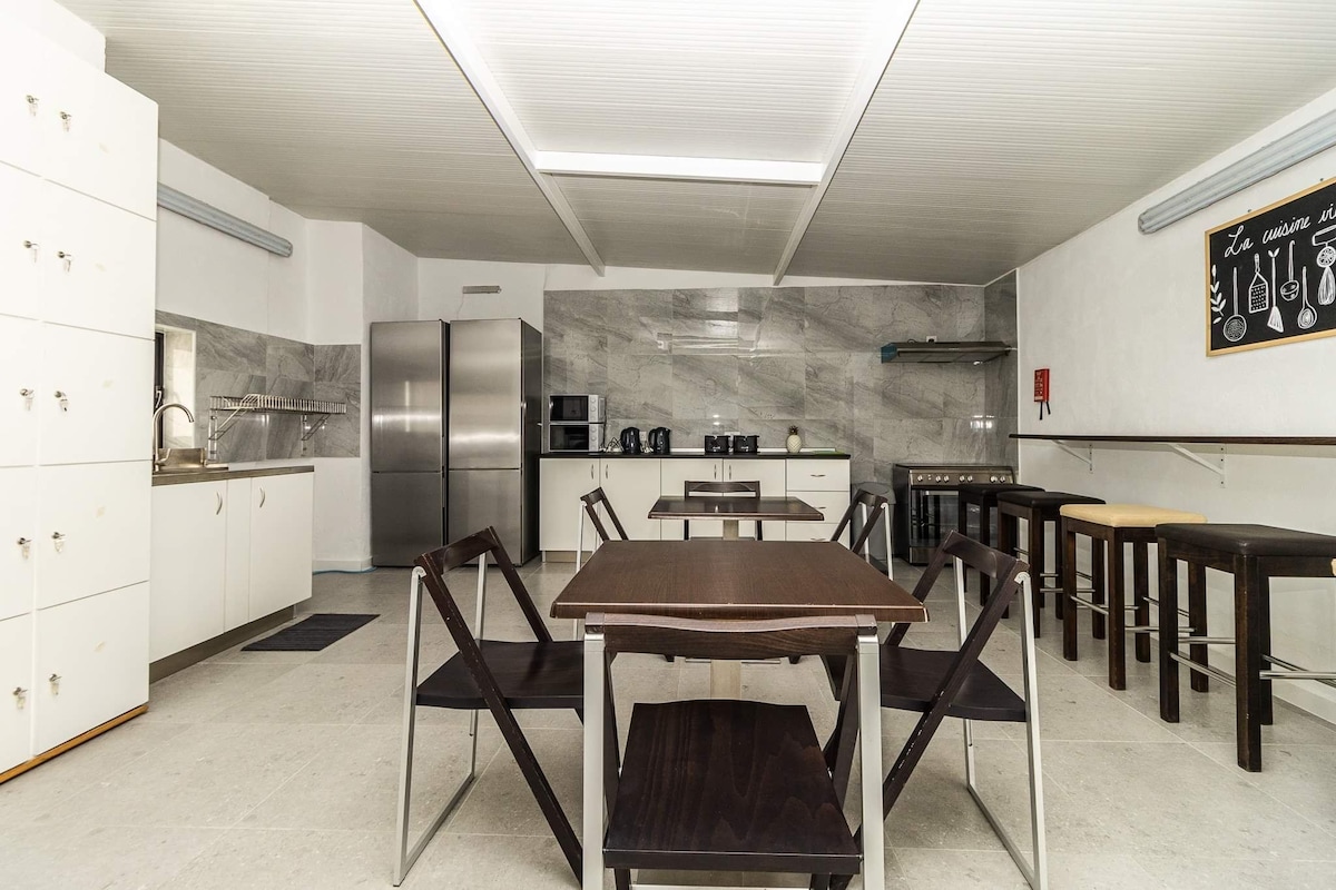 A spacious communal kitchen area features modern appliances, including a refrigerator, microwave, and coffee machine. A large table surrounded by chairs occupies the center, with additional bar stools along a countertop. Light-colored cabinets and a marble backsplash enhance the clean, functional design.