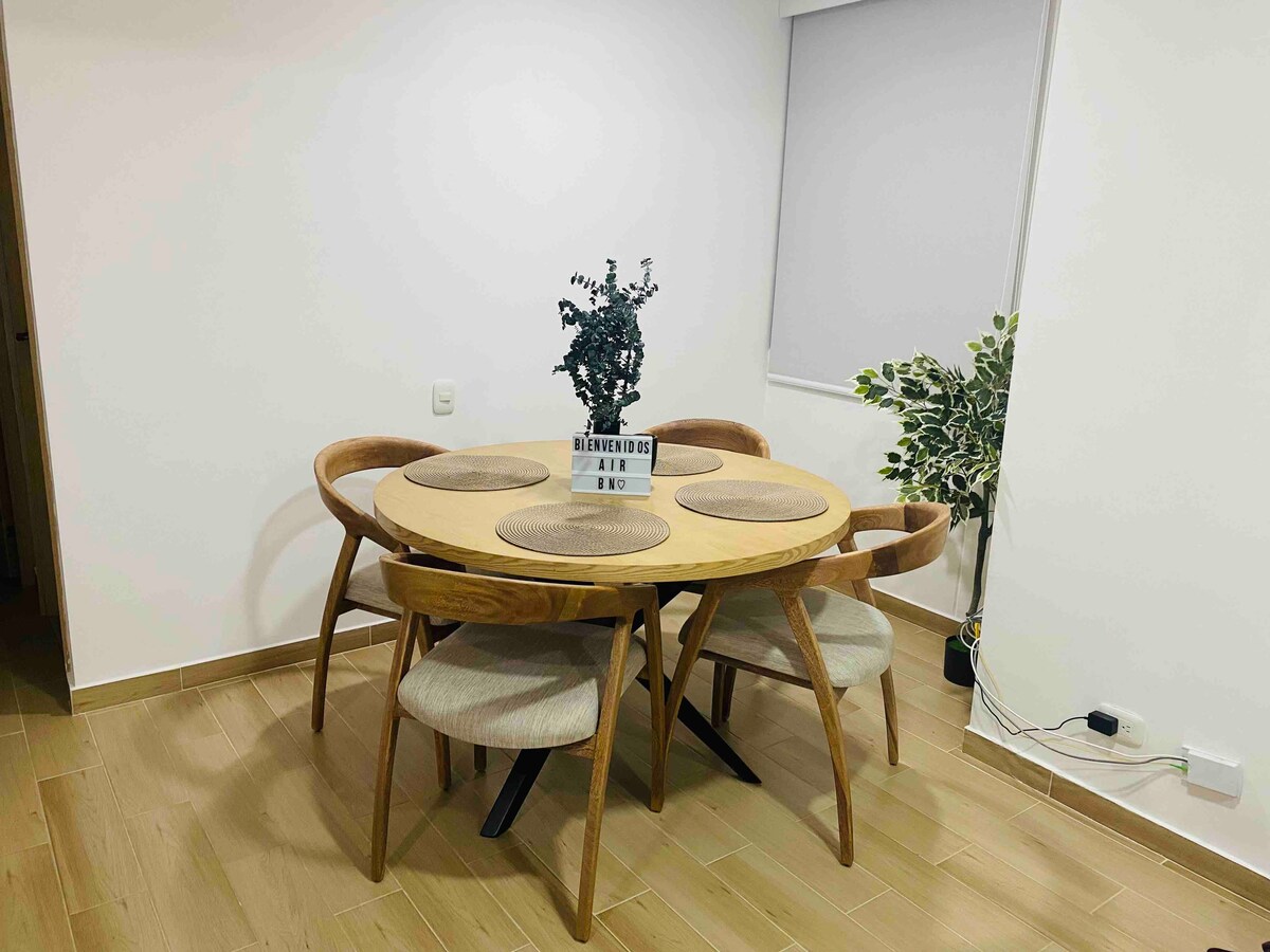 A round wooden dining table is surrounded by four curved-back chairs. Each chair features a cushioned seat. A small potted plant sits at the center of the table, while a plant is positioned against the wall in the background. Natural light enters through a window covered by a light blind.