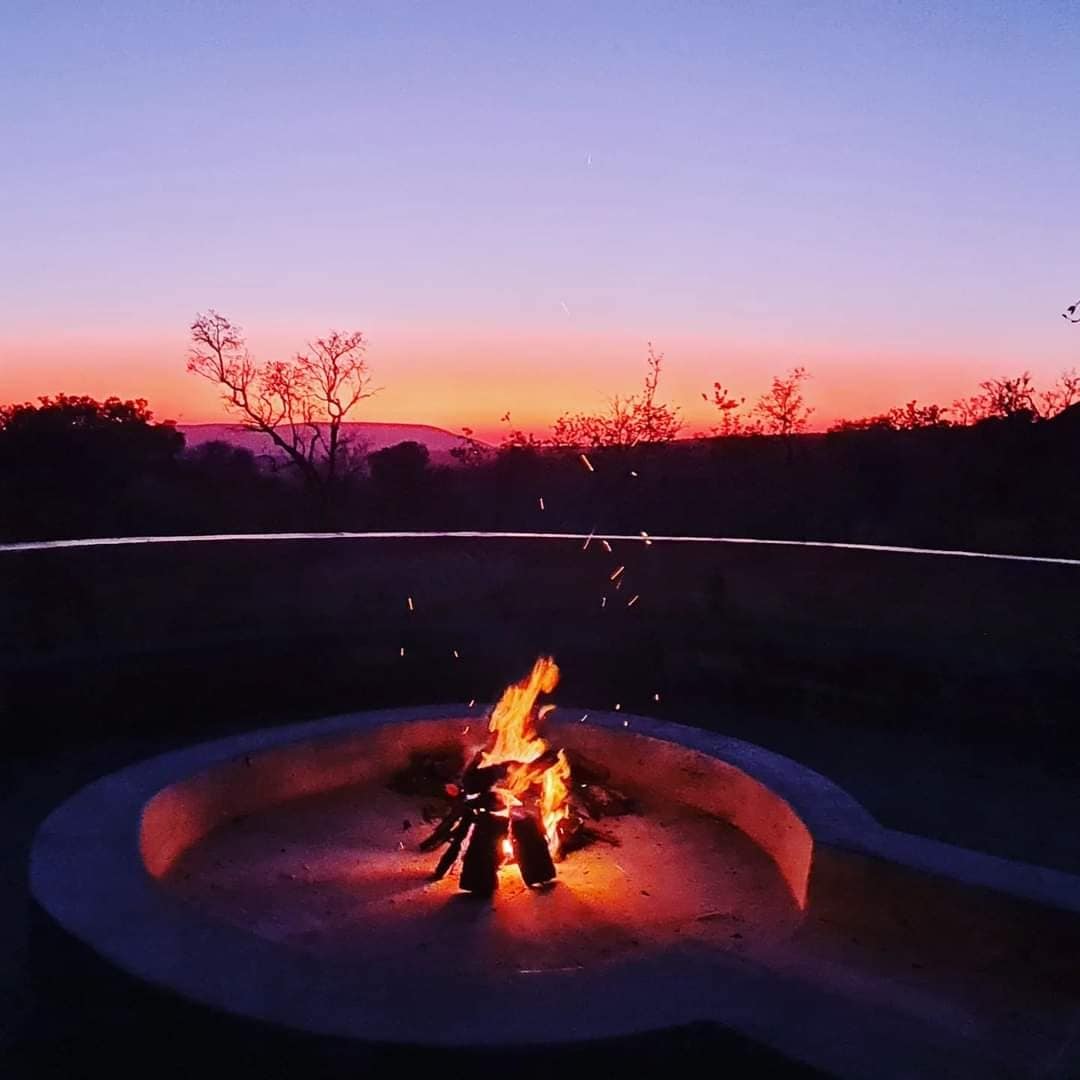 A circular fire pit is centered in the image, with flames rising and glowing embers spreading outward. A gradient of colors in the sky transitions from deep purple to soft orange and pink, signaling sunset. Silhouetted trees add depth to the serene landscape.