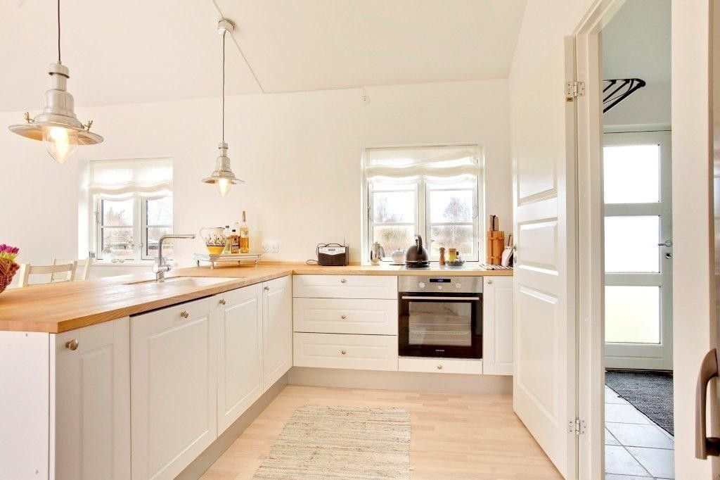 A bright kitchen features white cabinetry and natural wood countertops. Modern appliances, including an oven and stovetop, are visible. Two windows allow natural light, with a small area rug enhancing the space. Open shelves display kitchen essentials, contributing to an organized atmosphere.