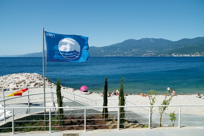 A clear view of a beach features a gently waving blue flag denoting quality standards. Sunbathers are seen on a pebbled shore, with lush greenery along the walkway leading to the beach. Mountains are visible in the background, complementing the serene coastal scenery.