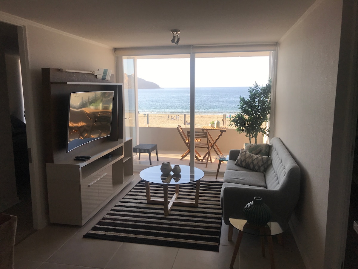 A bright living area is showcased with large glass doors providing a view of the ocean. A gray sofa is positioned near a coffee table on a striped rug, and a flat-screen television is mounted on the wall. A small dining set and a decorative plant complement the space.
