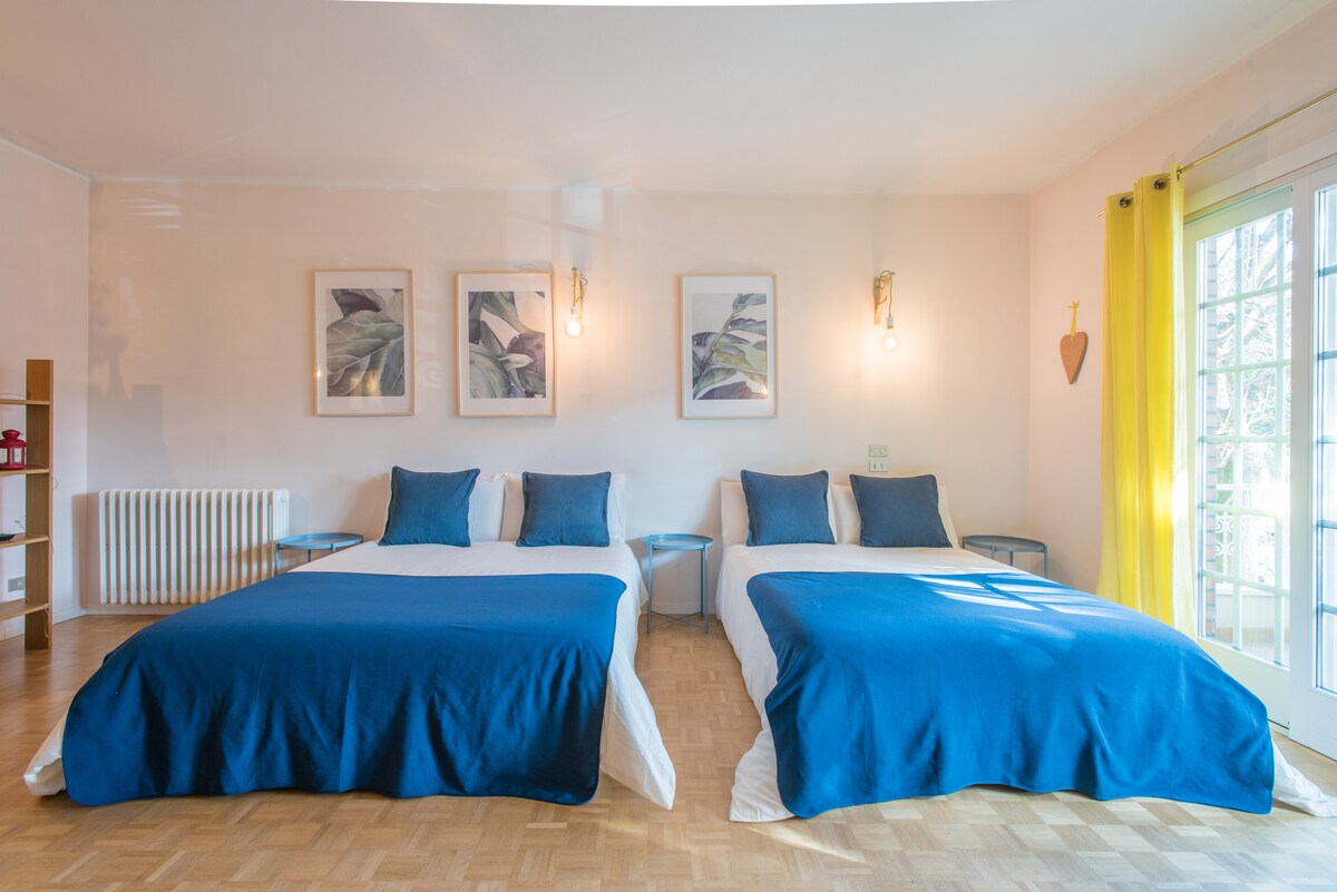 A spacious bedroom features two large beds dressed in white linens with deep blue throws. Framed botanical prints adorn the walls, and natural light enters through glass doors leading to a terrace. Bright yellow curtains add a cheerful accent to the room.
