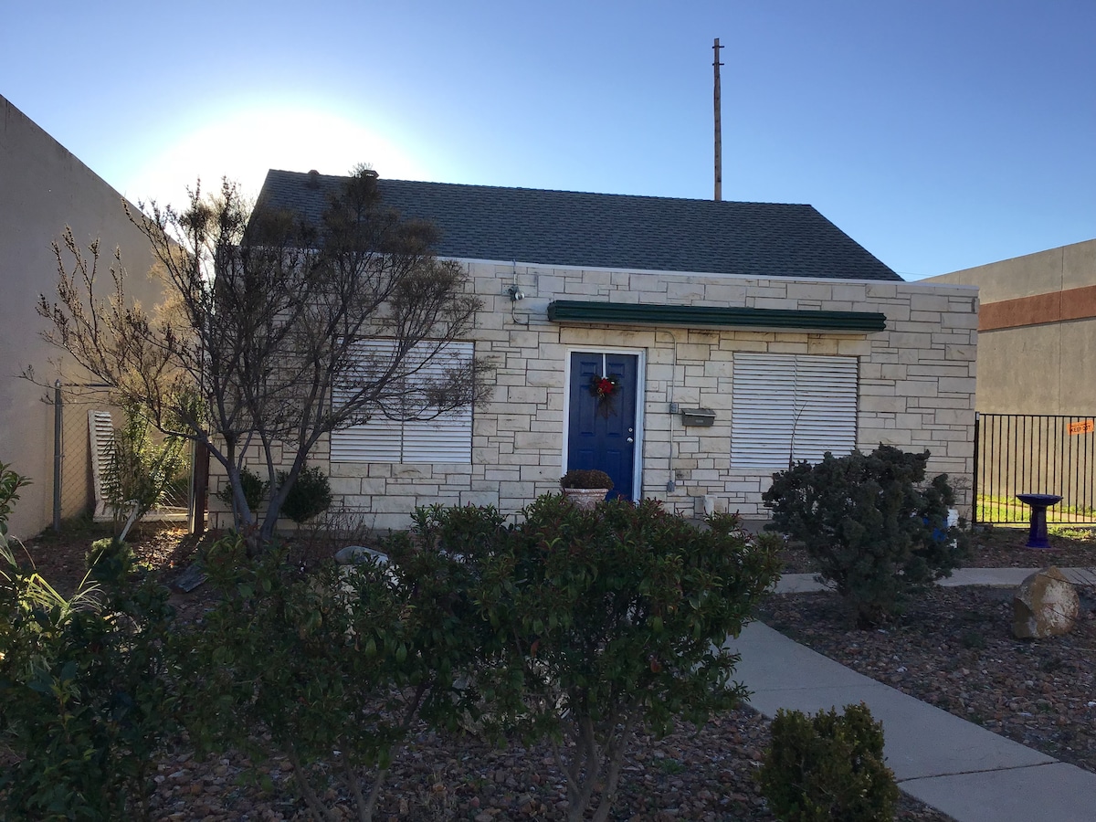 The cottage features a stone exterior with exterior shutters and a vibrant blue front door. Landscaping includes low shrubs and decorative stones that enhance the entrance. Visible behind the cottage are business buildings, illustrating the urban setting of the property.