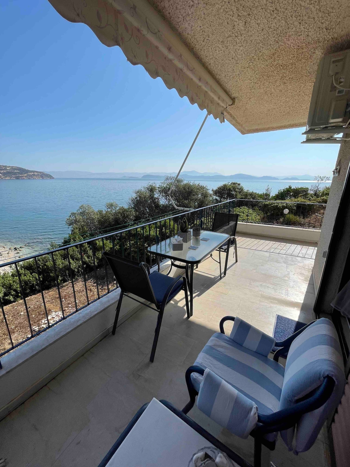 A balcony overlooks the ocean, featuring a table with two chairs positioned to enjoy the view. Soft sunlight illuminates the space, and greenery is visible along the shoreline. The tranquil water reflects the clear blue sky, enhancing the peaceful atmosphere.
