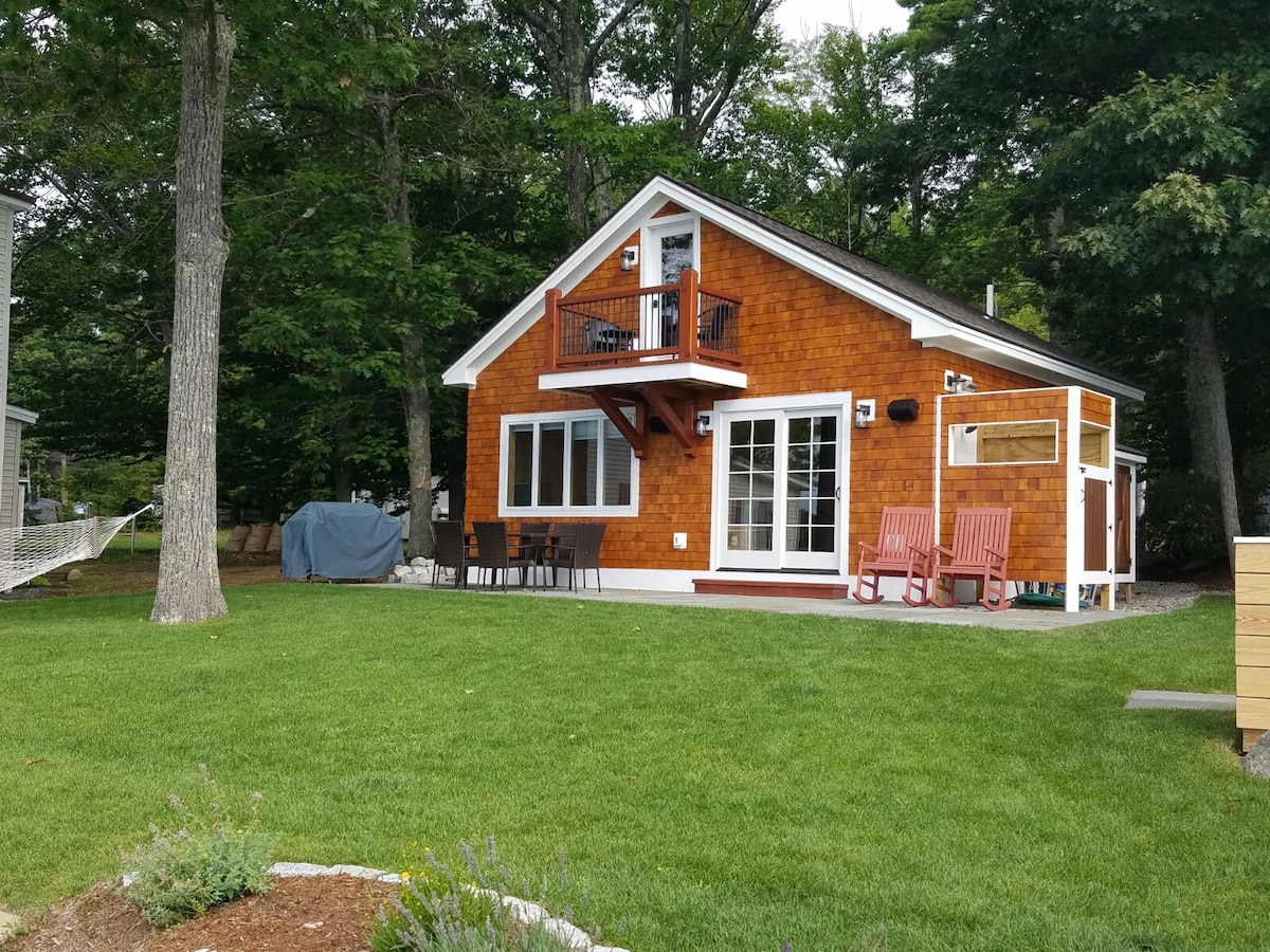 A charming lakeside cabin features a wooden exterior with white trim. Large windows allow natural light to fill the open concept living area. A private balcony overlooks the backyard, which is adorned with a well-maintained lawn and seating area, surrounded by tall trees.