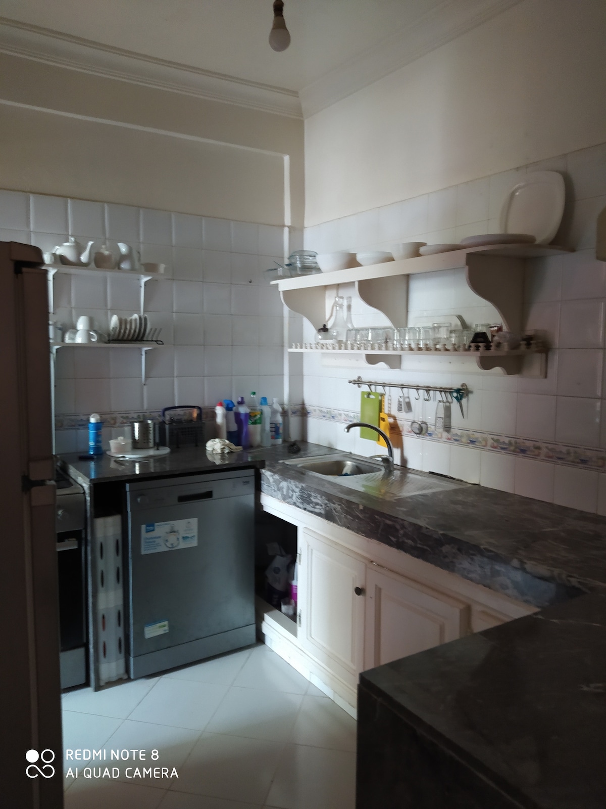 The kitchen features a marble countertop with a sink and essential appliances visible, including a refrigerator and dishwasher. Shelves hold various dishware and glassware, while cleaning supplies are organized beneath the counter, ensuring a functional and tidy workspace.
