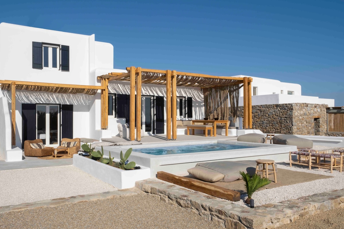 The villa's exterior showcases a modern design with wooden beams providing shade over the spacious pool area. Comfortable loungers are arranged around the pool, surrounded by minimalist landscaping, while the white walls reflect the bright blue sky.