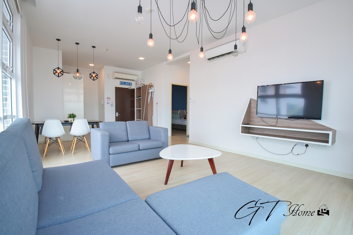 A modern living space features a light-blue sofa and a white coffee table. Two dining chairs accompany a round dining table, while soft lighting is provided by pendant lamps. A wall-mounted television is positioned opposite the seating area, adding functionality to the room.