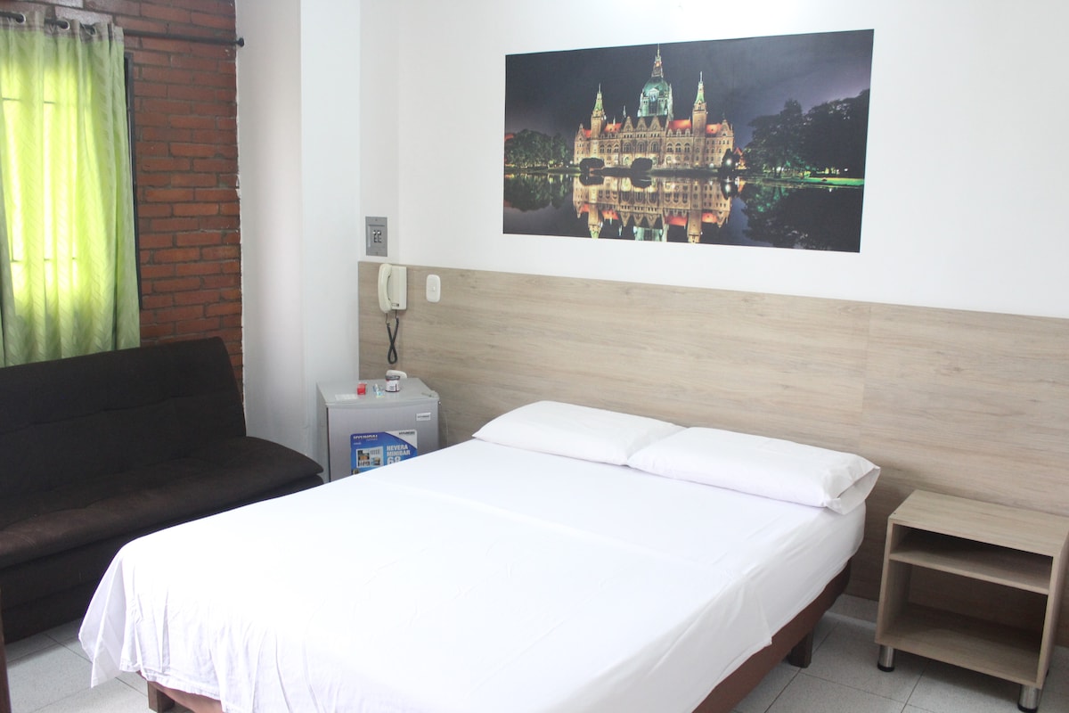 A comfortable room is presented with a spacious bed adorned with crisp white linens. Adjacent, a dark brown sofa adds seating. A striking wall mural displays a cityscape, while a window draped with light green curtains allows natural light to enter, complementing the neutral decor.