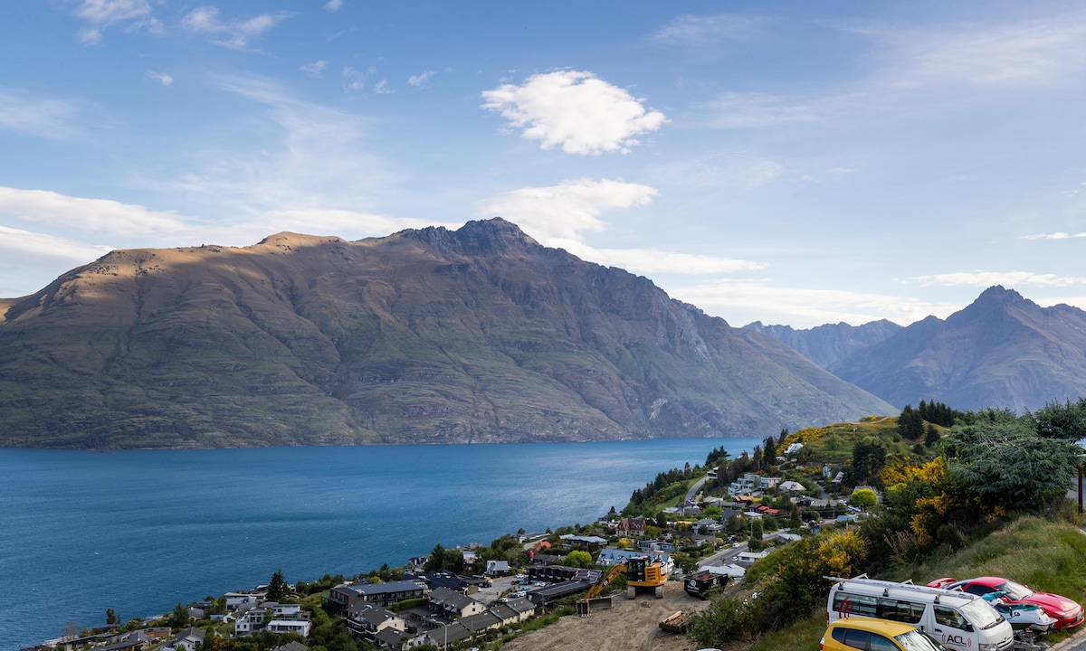 A panoramic view of Lake Wakatipu, reflecting the sky and surrounded by mountainous terrain. The scene captures lush greenery and a residential area below, with various vehicles parked along the road, showcasing the natural beauty of the landscape.