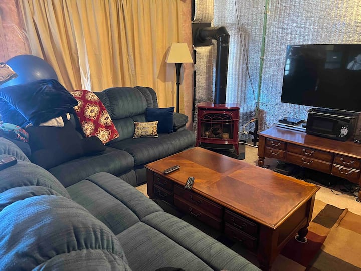 Eneo la kukaa la chini ya ardhi ambalo halijakamilika, lenye kipasha joto cha propani cha jiko la mbao kutoka Vermont Castings maarufu ulimwenguni na televisheni mahiri.  Sofa iliyo karibu zaidi na jiko ni kitanda cha sofa.