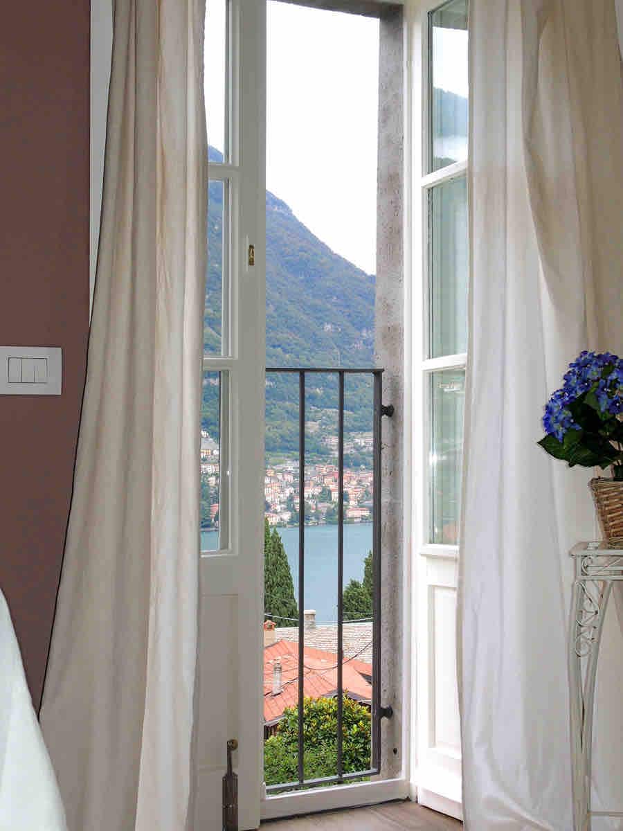 A window with white curtains frames a view of the lake and surrounding mountains. A small balcony with a metal railing is visible, and a flower pot sits on the windowsill, adding a touch of greenery to the serene setting.