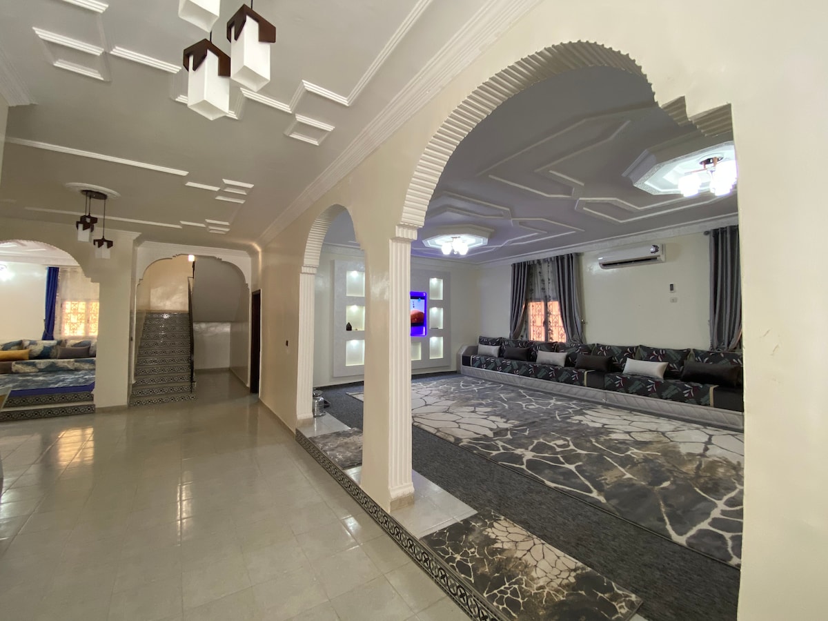 A spacious living area is highlighted by soft lighting and decorative arches. A large plush sofa is arranged around a textured area rug, while an entertainment unit is positioned against the wall. Floors are finished with neutral tiles, providing a clean and open feel.