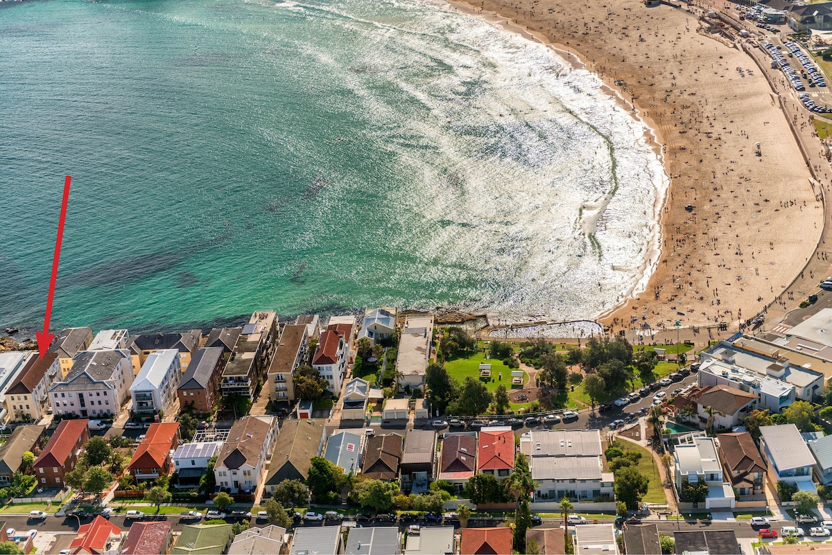 An aerial view highlights the apartment's proximity to Bondi Beach, with the coastline clearly visible. The surrounding neighborhood features a mix of houses and greenery, while the sandy beach is populated with beachgoers enjoying the sun and surf.