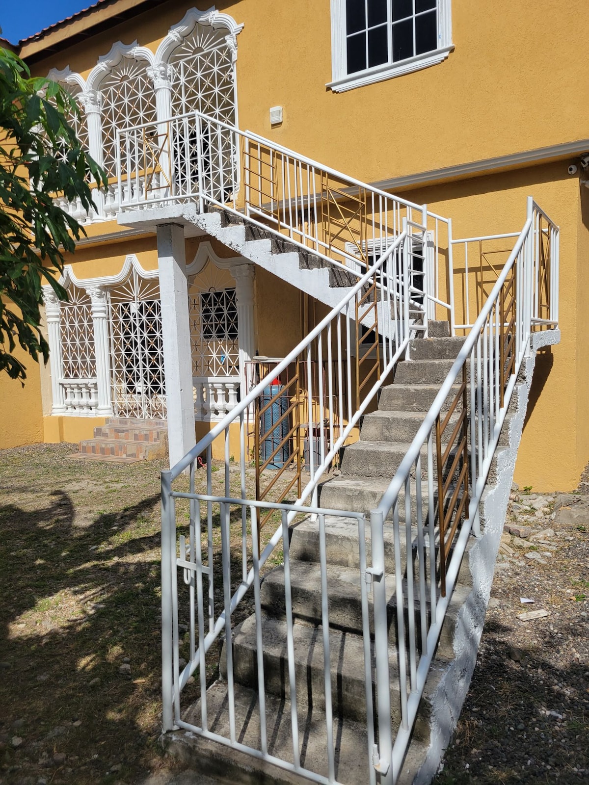 A set of white metal stairs is seen leading up to a brightly colored exterior. Decorative railings frame the stairs, providing safety. A large window with intricate metalwork is visible next to the stairway, showcasing architectural details of the building.