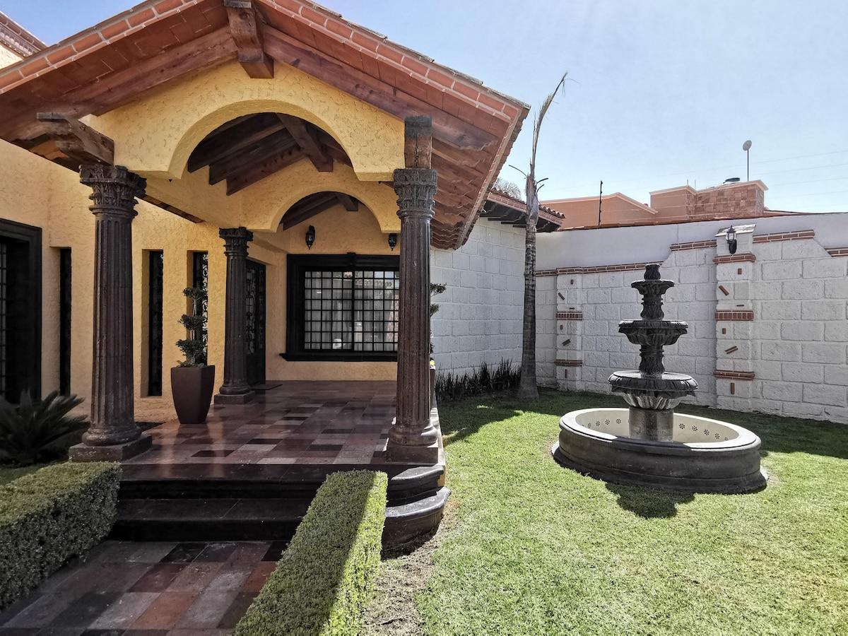 An outdoor area features a well-maintained lawn bordered by shrubbery, with a decorative fountain at the center. The entrance is marked by a covered porch supported by distinct columns, showcasing traditional architectural elements. A large window provides visibility into the interior space.