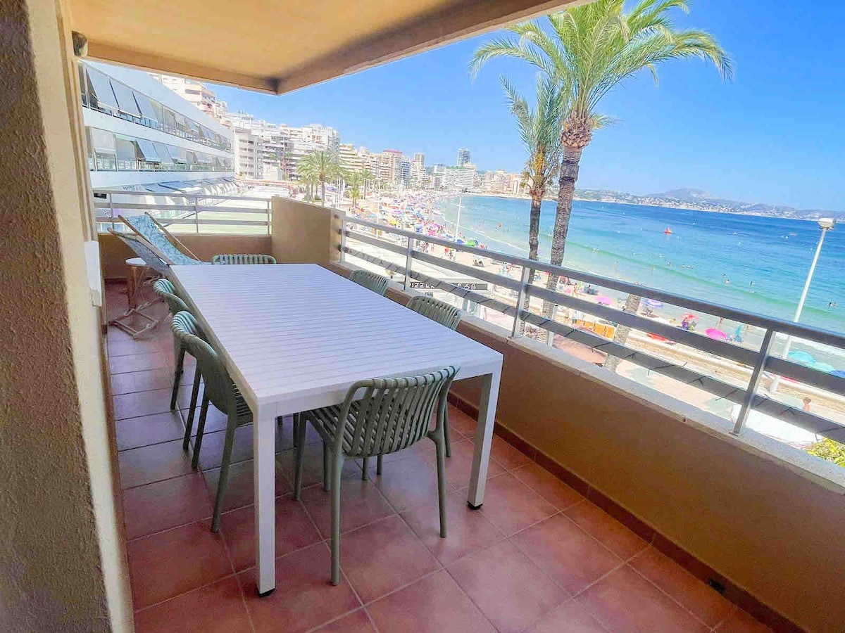 A dining table with several chairs is positioned on a balcony overlooking a scenic beach view. Palm trees are visible along the shoreline, and the turquoise water stretches toward the horizon. The beach area is populated with loungers and colorful umbrellas.