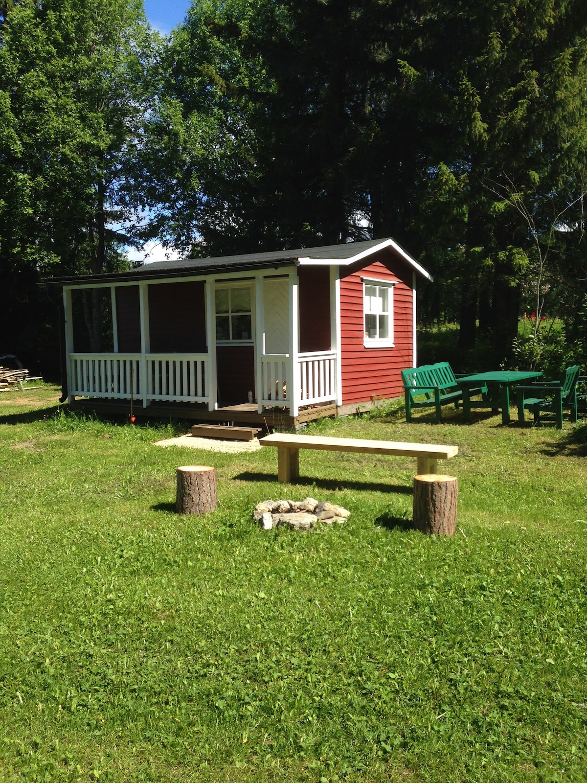 A red cottage with white trim features a front porch and large windows. A wooden bench made from logs is positioned near a fire pit, surrounded by a grassy area. Outdoor seating is visible, inviting relaxation in the natural surroundings.