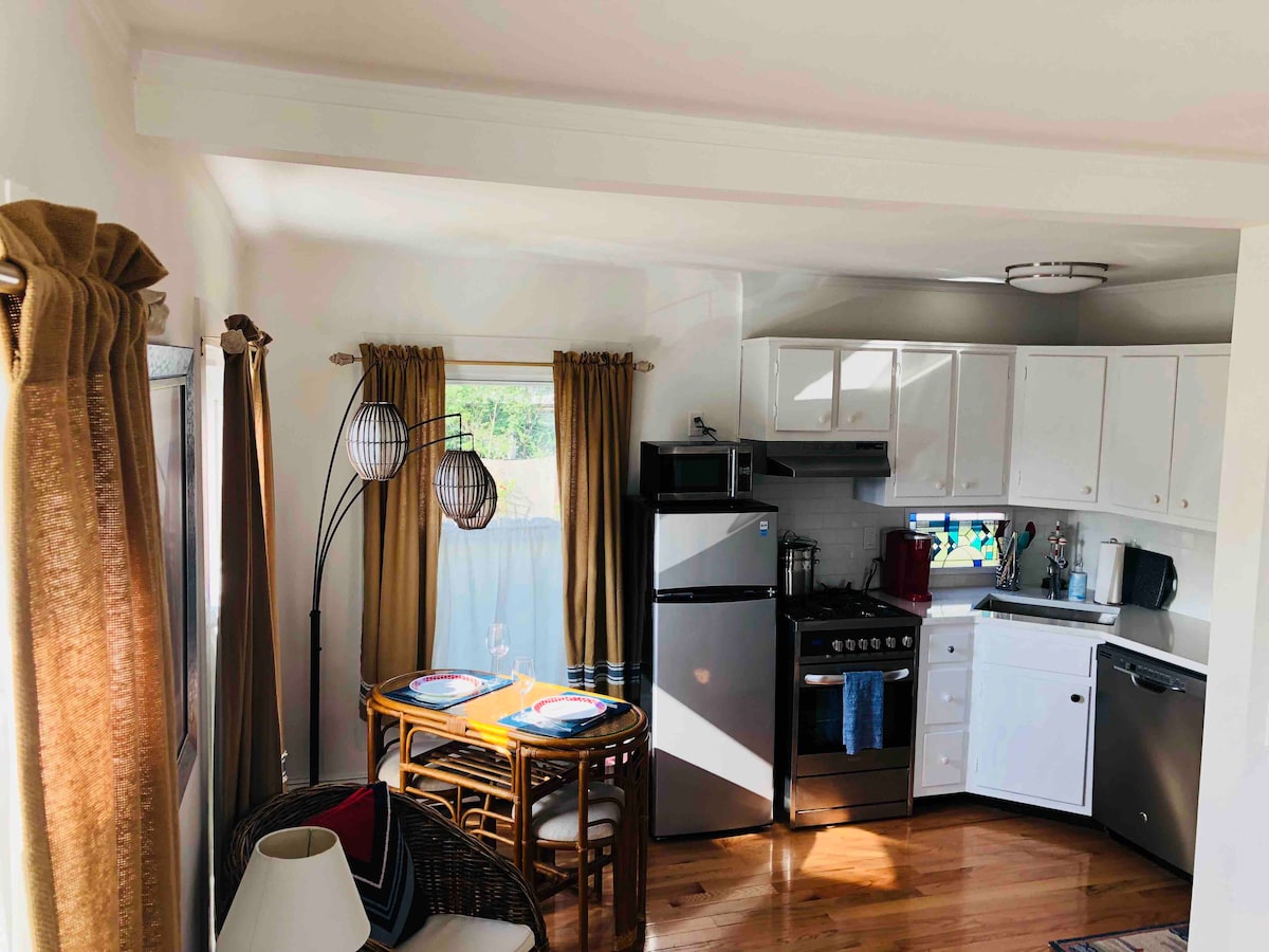 A bright kitchen area features white cabinetry and stainless steel appliances. A dining table for two is set with colorful plates, enhanced by natural light streaming through the window. Soft golden curtains frame the surroundings, contributing to the warm atmosphere of the space.