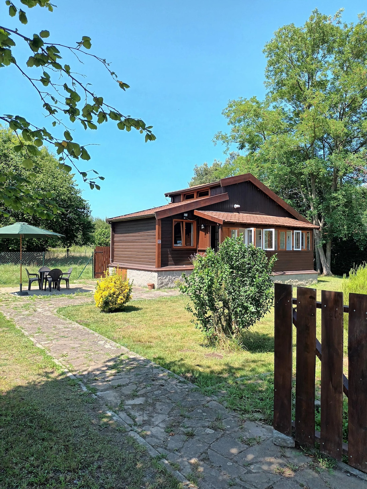 A charming wooden cabin is surrounded by green lawns and trees. A stone path leads through the garden, which features a small outdoor dining area under a green umbrella. The structure has multiple windows, allowing for ample natural light.