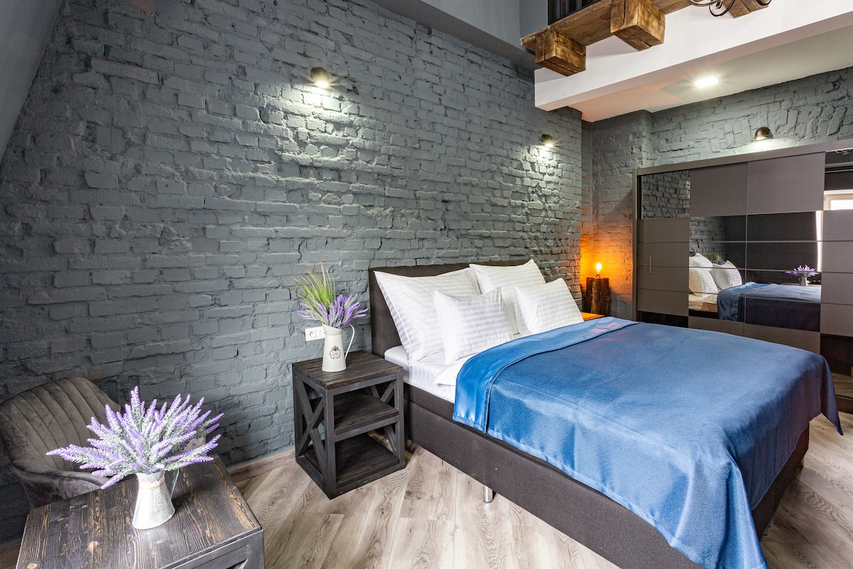 A welcoming bedroom is enhanced by gray brick walls and warm lighting. A spacious bed is dressed in blue linens, with white pillows arranged neatly. A small bedside table holds a vase with lavender, while a modern wardrobe reflects the room's contemporary design.