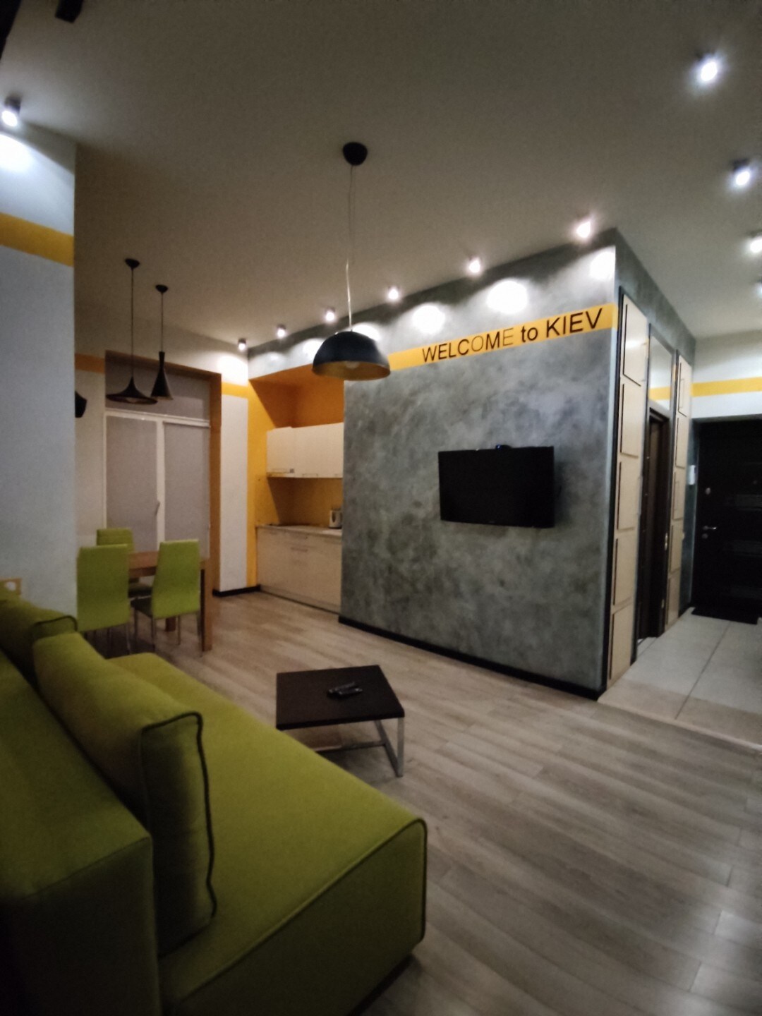 The living area features a green sofa with a dark coffee table, offering a cozy spot. A dining table with green chairs sits adjacent to a compact kitchen with modern appliances. A flat-screen TV is mounted on a well-lit, textured wall that includes a welcoming message.