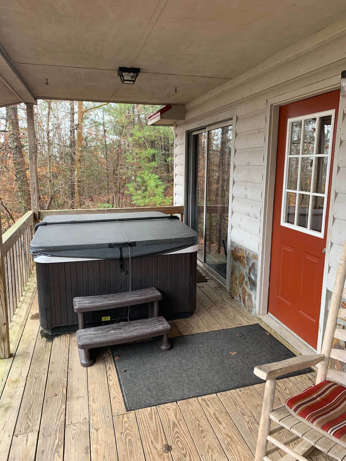 A hot tub is situated on a wooden porch, accompanied by a set of steps for easy access. The entrance features a bright orange door, leading into the cabin. A rocking chair is positioned near the railing, with views of the surrounding trees.