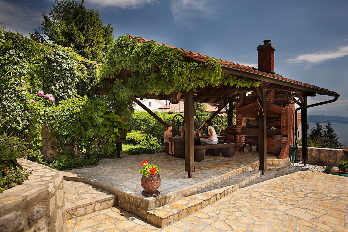 An outdoor dining area is sheltered by a wooden pergola adorned with lush greenery. Stone flooring leads to a barbecue structure, with a large table and bench seating available under the shaded space. A peaceful landscape and serene sky enhance the setting.