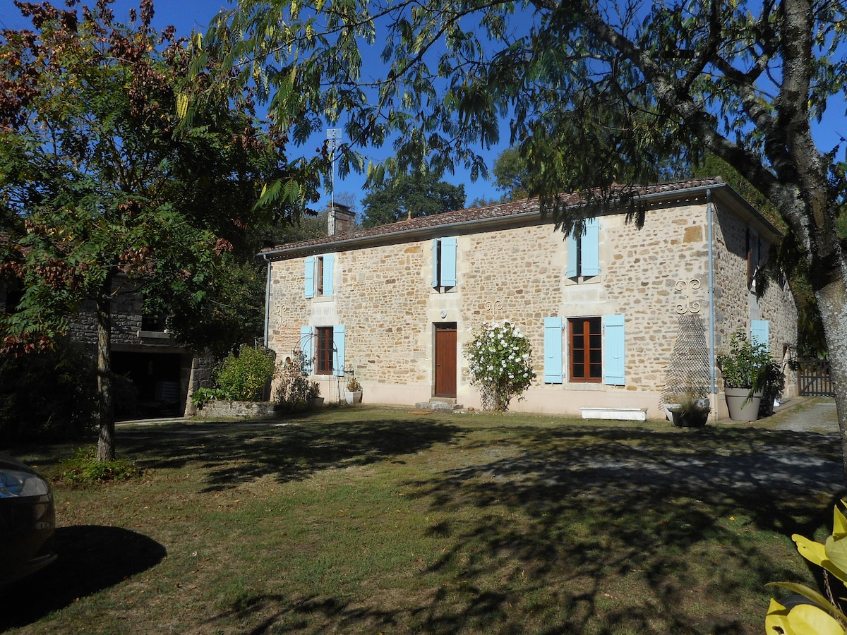 A characterful stone house is surrounded by greenery, featuring light blue window shutters and a welcoming front door. Lush grass and trees create a serene atmosphere, while the structure's heritage style reflects its historical charm.