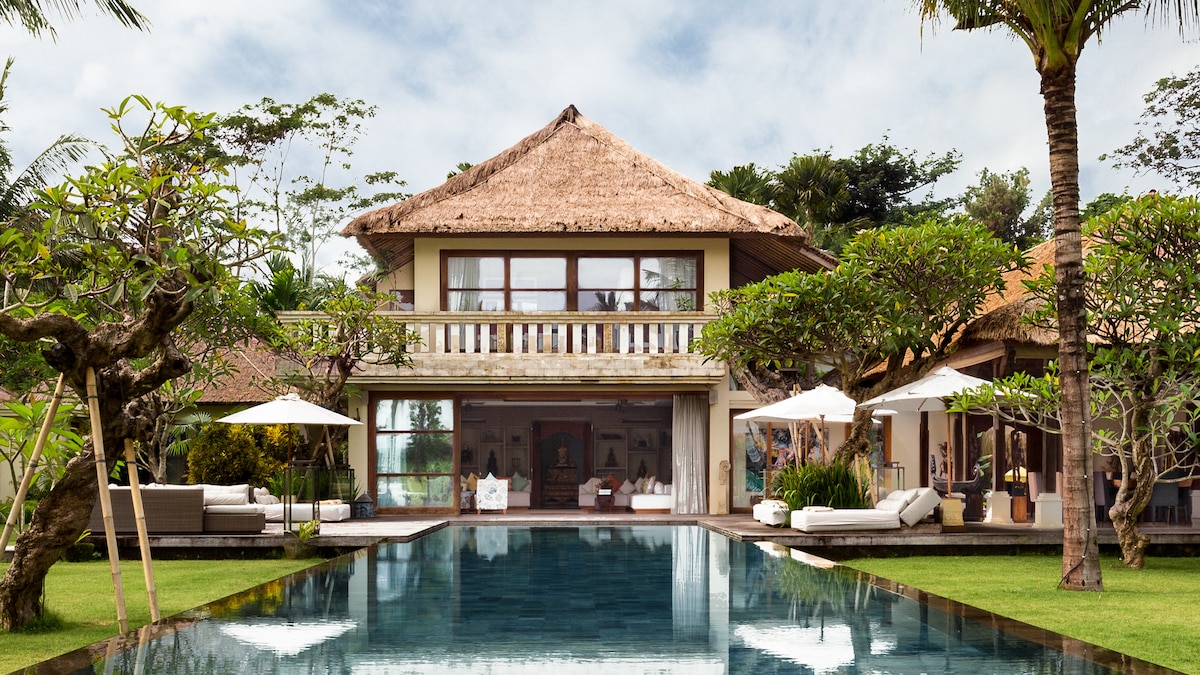 The spacious estate features a thatched roof with expansive glass walls, allowing for natural light. Lush tropical greenery surrounds a tranquil reflecting pool, bordered by sun loungers and umbrellas. Manicured lawns create a serene atmosphere, inviting relaxation in the outdoors.